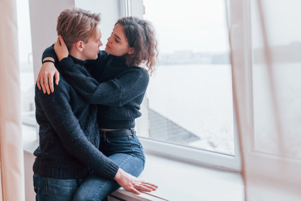 一对可爱的年轻夫妇在室内靠近窗户的地方拥抱在一起图片下载