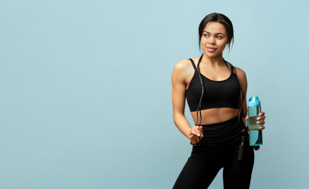 快乐的非洲裔美国女士跳绳和淡水瓶在蓝色背景与空白摆姿势图片下载