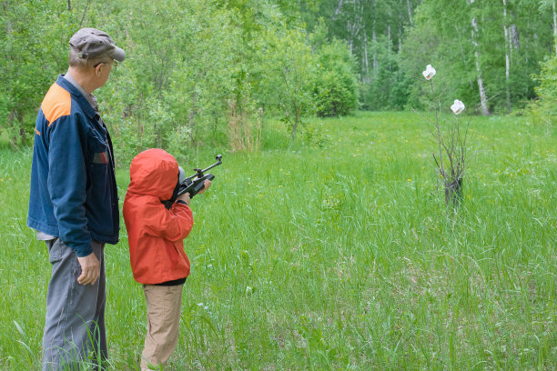 一个穿着红色风衣的男孩的后视图是瞄准气枪与一个戴着帽子的祖父在夏季公园。概念武器和孩子，尊重，小心，训练。图片下载