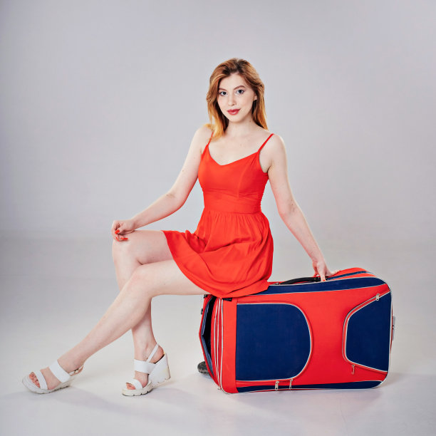 棕色头发的女孩穿着红色的裙子坐在一个大的红蓝色的手提箱在灰色的背景。图片下载