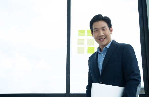 亚洲商人站在办公室，手里拿着笔记本电脑，自信地微笑。图片下载