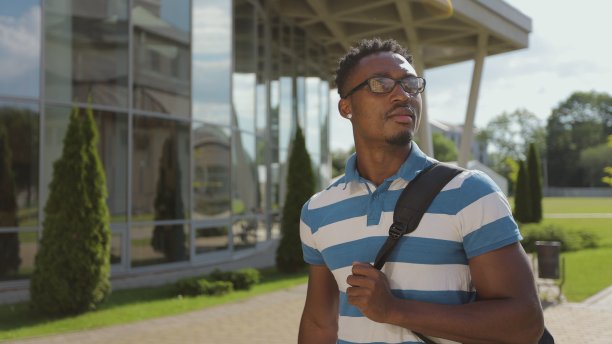 非洲裔男学生背包行走在大学校园。一个年轻的美国黑人戴着眼镜，背着背包走在现代城市的街道上。图片下载