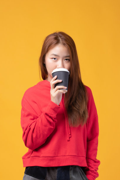 一个漂亮的年轻亚洲女人的肖像，微笑着拿着外卖咖啡杯穿着红色毛衣孤立在黄色的背景。美丽的年轻女孩喜欢在早晨喝咖啡。图片下载