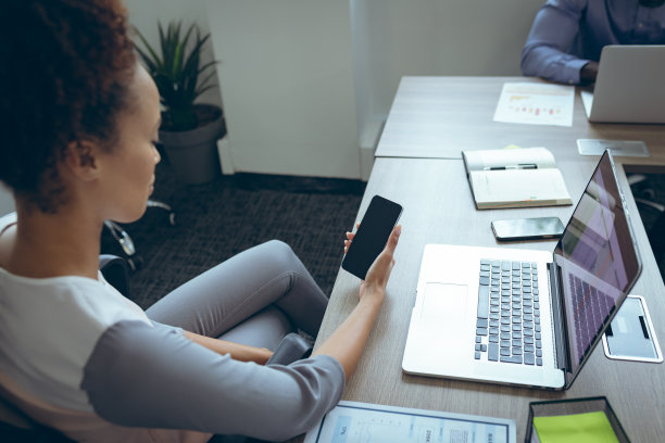 混血女商人坐在办公桌前使用智能手机，同事在后面图片下载