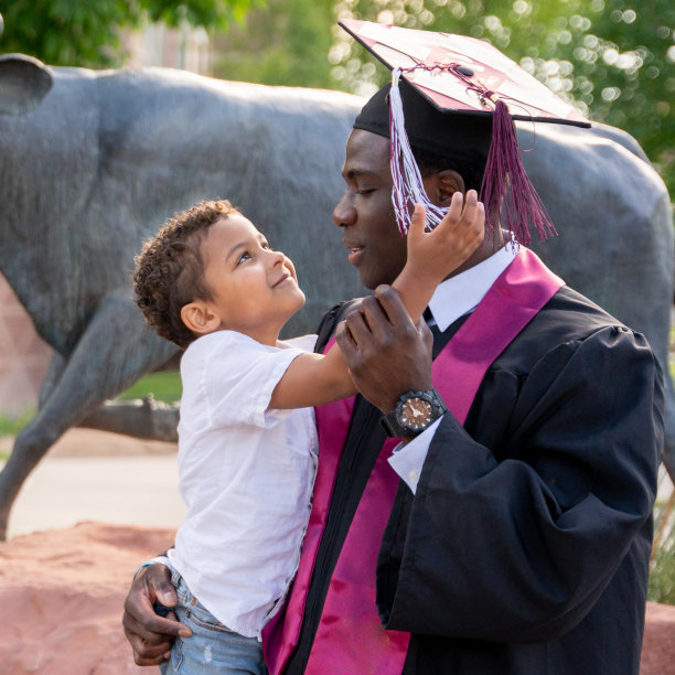 非裔美国男大学毕业生戴着帽子和长袍，在毕业典礼上和他三岁的儿子玩图片下载