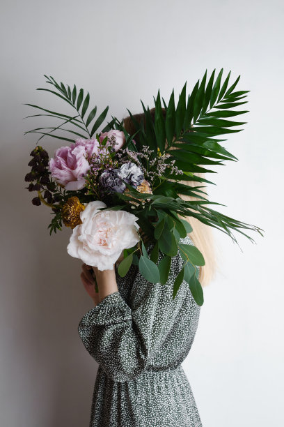 一束丰富的牡丹，热带树叶和美丽的不同的花朵在一个女孩的手在一个明亮的背景。卖花的女孩收集了一束花。浪漫喜庆的花束。节日礼物。图片下载