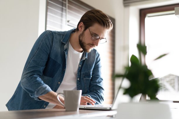 年轻人随便上网用笔记本电脑工作，站在厨房桌子旁边，看着电脑屏幕图片下载