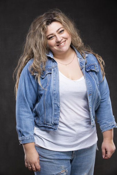 一个穿着牛仔夹克的漂亮胖女人看着镜头，在灰色的背景上微笑着。图片下载