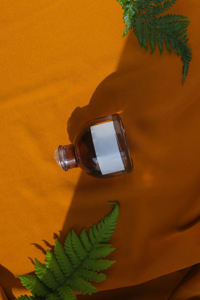 香水瓶与空白白色标签和绿色叶子在纹理的橙色织物背景。香水瓶库存照片图片下载