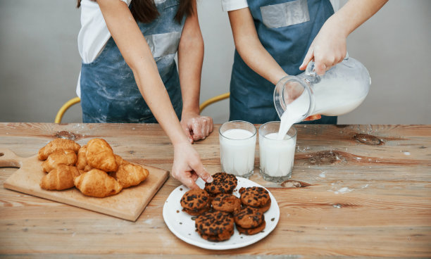 近距离观看两个小女孩在蓝色厨师制服，倒牛奶到杯子上的厨房和桌子上的饼干图片下载
