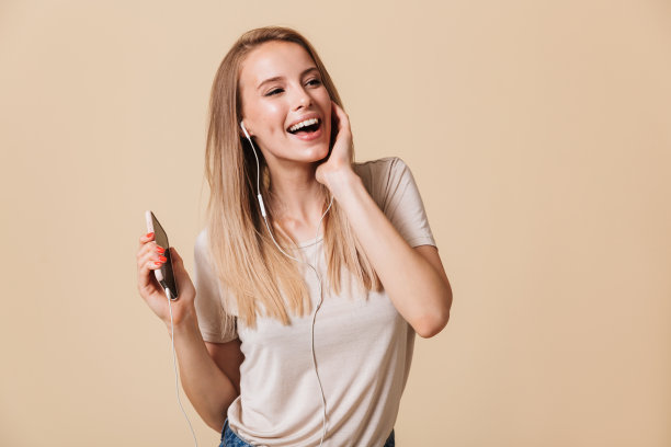 一个开朗的金发女人穿着休闲衣服一边唱歌一边通过耳机听着智能手机上的音乐，在录音室里被米色的背景隔开图片下载