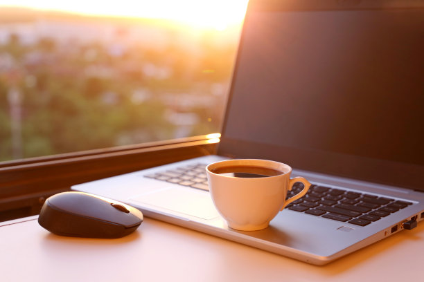 日落时分，热咖啡杯和笔记本电脑放在靠窗的桌上图片下载
