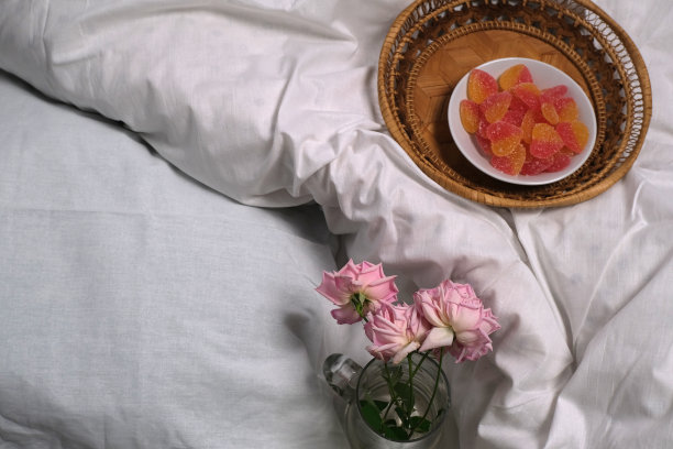 精致的粉红玫瑰，笔记本，果酱放在一张白色的未整理的床上。图片下载