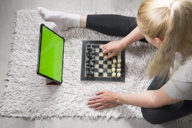 一个女人在地毯上和一个虚拟的伴侣在平板电脑上下棋，平板电脑有一个绿色的屏幕。远程在线游戏图片下载