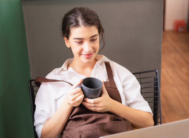 美丽的白人女咖啡师持有热咖啡杯在咖啡休息，同时使用笔记本电脑在咖啡店图片下载