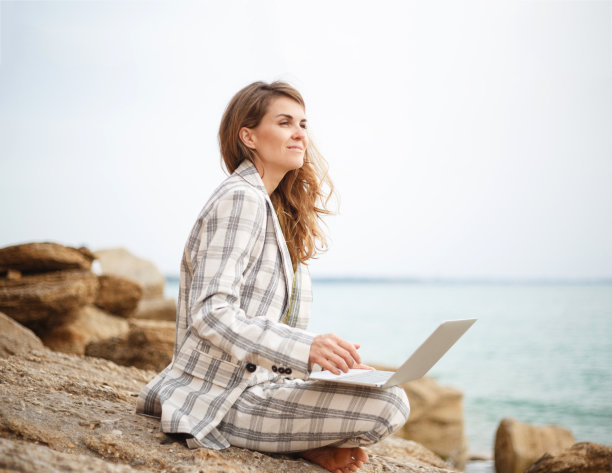 一个年轻女子坐在海边的一块石头上，手里拿着笔记本电脑。远程工作的概念图片下载