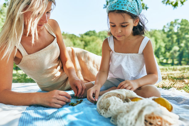 慈母和她可爱的女儿坐在毯子上玩耍，在一个阳光明媚的夏天在公园里探索。女人和她的孩子有舒适的夏天野餐与柠檬环保袋。母亲节概念。图片下载