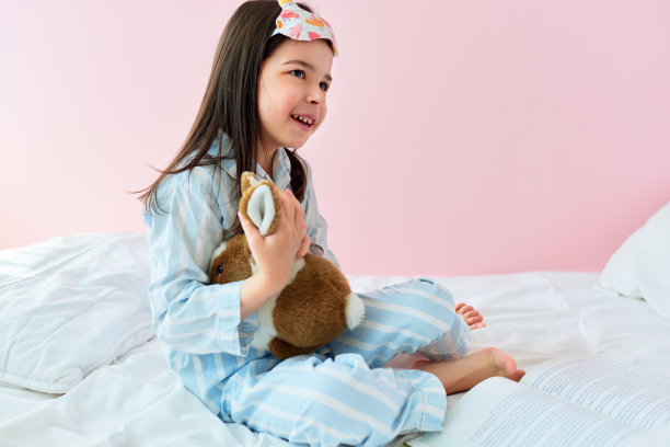 一个顽皮的小女孩穿着睡衣在床上玩玩具在现代舒适的阳光卧室的侧视图。快乐的孩子休息在床上在早上在家里在她的生日。图片下载