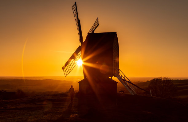 日落时，太阳在一个传统风车后面的地平线上落下，画面上是一个人在遛狗图片下载