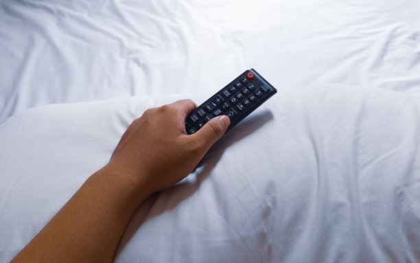 亚洲男子手握电视遥控器上方白色枕头和床图片下载