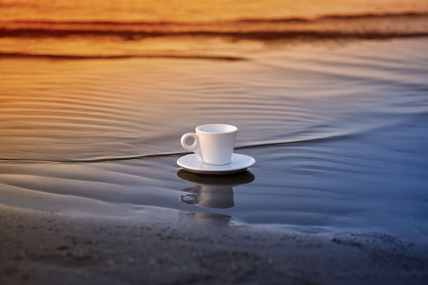 沙滩上的白色咖啡杯与温暖的阳光-选择性焦点。海滩上的咖啡杯。早上在海滩上喝咖啡。图片下载