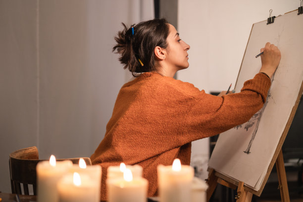 一位用铅笔把棕色长发盘成一个髻的妇女晚上坐在蜡烛旁。一个专注的女孩带着艺术家的灵魂画了一幅树的画，在一个洞穴上素描图片下载