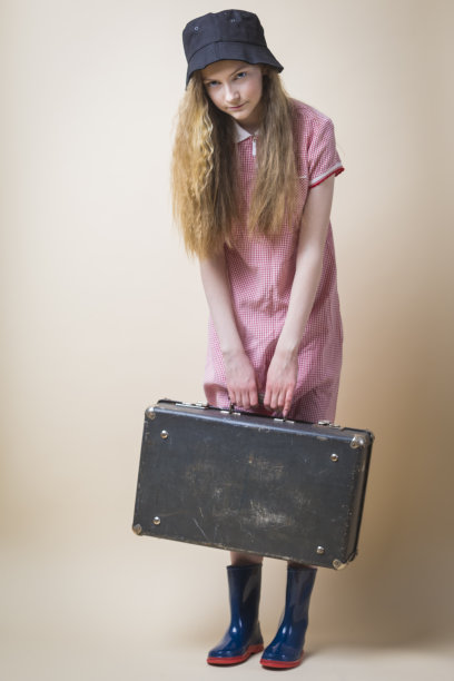 疲惫的白人少女穿着红色格子连衣裙和威灵顿靴，头戴帽子，手提旧旅行箱，竖版照片图片下载