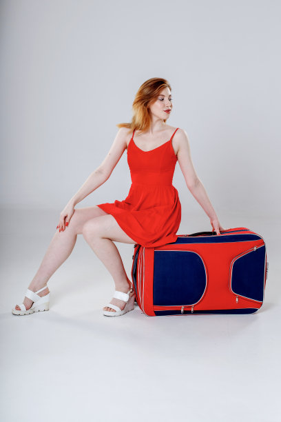 一位棕色头发、身穿黑色连衣裙的妇女坐在灰色背景上的一个红蓝相间的大手提箱上。图片下载