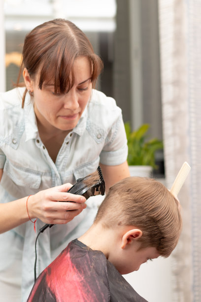 在隔离期间，年轻的理发师妈妈在家用理发器剪她的男婴。有选择性的重点图片下载