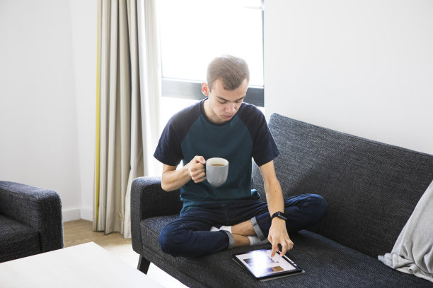 一名年轻的男学生盘腿坐在灰色的沙发上，手里拿着一杯咖啡，低头看电视。他穿着一件蓝绿色的t恤和牛仔牛仔裤。图片下载