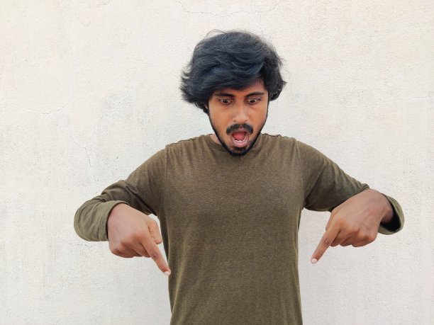 长头发的南印度年轻人向下看，手指向下，令人震惊的反应。打开嘴广泛。白色背景图片下载