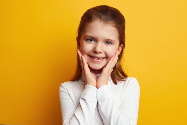年轻可爱的小女孩双手贴在脸颊上，对着黄色的墙壁微笑图片下载
