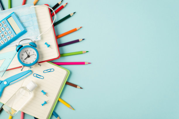 俯视图照片的多色铅笔，闹钟，尺子，计算器，剪刀，医用口罩，消毒剂，针，夹在打开的红色和绿色的抄写本孤立的彩色蓝色背景与抄写空间图片下载