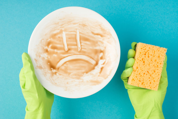 俯视图照片的手在绿色橡胶手套拿着白色的脏盘子与绘制悲伤的脸和黄色海绵在孤立的柔和的蓝色背景图片下载
