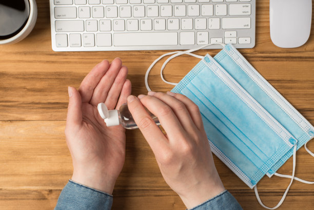 第一个人俯视图照片的手使用杀菌剂，透明的瓶子，两个医疗面罩，咖啡杯，键盘和鼠标在孤立的木桌背景与拷贝空间图片下载