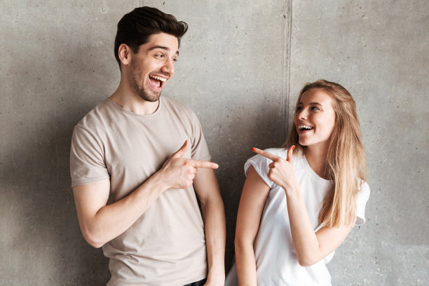 年轻人的肖像，20多岁的男孩和女孩穿着基本的衣服微笑着互相指着对方，孤立在室内的混凝土灰色墙壁上图片下载
