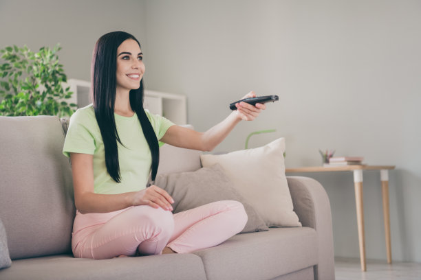 迷人的女孩的照片坐在沙发，手持遥控器，交叉的双腿，闪亮的微笑，看穿绿色的t恤，粉红色的裤子在室内图片下载