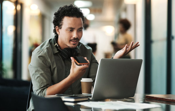 一个商人坐在办公室里用他的手提电脑用他的手机发送语音提示的照片图片下载