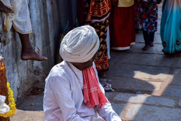 这是印度卡纳塔克邦Yadgir的一张照片，50到60岁的印度老人穿着传统的白色衣服和白色头巾，独自坐在印度教寺庙的楼梯上，阳光灿烂。图片下载