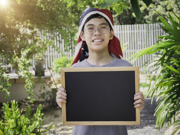 小男孩戴着美国国旗头巾，举着空的或空白的标志，白色的黑板和站在花园里。退伍军人或独立日的概念。图片下载