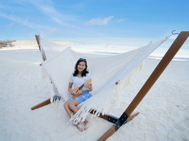 亚洲妇女坐在白色沙滩上的白色吊床上弹奏尤克里里琴图片下载