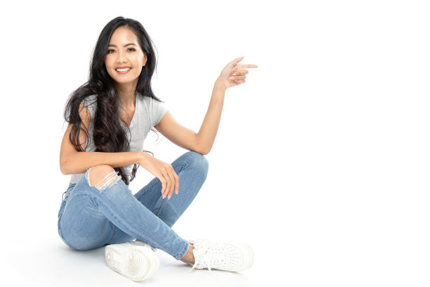 一位穿着休闲装的年轻亚洲女子坐在地板上。她用手指着旁边的复印版面。孤立在白色背景上。在白色背景上隔离图片下载