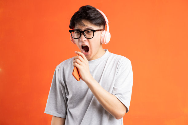 一名年轻的亚洲男子戴着耳机拿着智能手机唱歌的照片，在橙色背景下被孤立图片下载