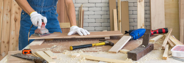 木工在木工设施中使用砂光机用砂纸磨光胶合板表面。一张满是手工工具和木堆的桌子。图片下载