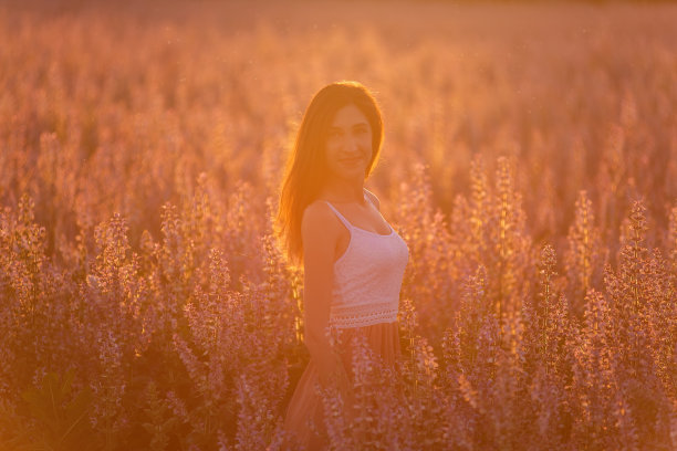 年轻女子在白色t恤，粉短裤站在盛开的粉红色鼠尾草田。照片中的女孩在橙色的阳光下，头发在日落的风中飘动。花纹理。出城旅行图片下载