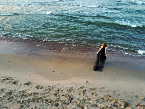 一位穿着黑色长裙的美丽女子站在海边的沙滩上。航拍的一个年轻的女孩时装模特休息在海洋上。穿着裙子和拖裾跳舞。海浪和生活的享受。图片下载
