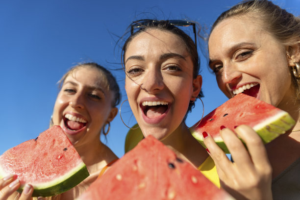 在夏天的时候，三个快乐的女孩在户外的海滩上吃西瓜。朋友们有有趣的孤立背景享受和咬新鲜的水果在海边度假。贞图片下载