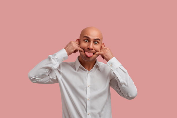 让乐趣。横向照片的快乐积极光头男子与刚毛，伸出舌头做疯狂的鬼脸，延伸一个微笑与她的手指。穿着白衬衫，在粉红色的背景下显得很孤立。图片下载