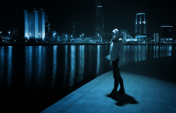 一个戴着防毒面具的女人的剪影，背景是一座夜城和一个池塘，这是冠状病毒大流行的启示概念。图片下载