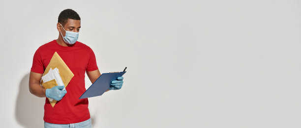 忙碌的快递员戴着口罩和防护手套看着剪贴板，拿着包裹和信封，在浅灰色的背景下摆出孤立的姿势图片下载
